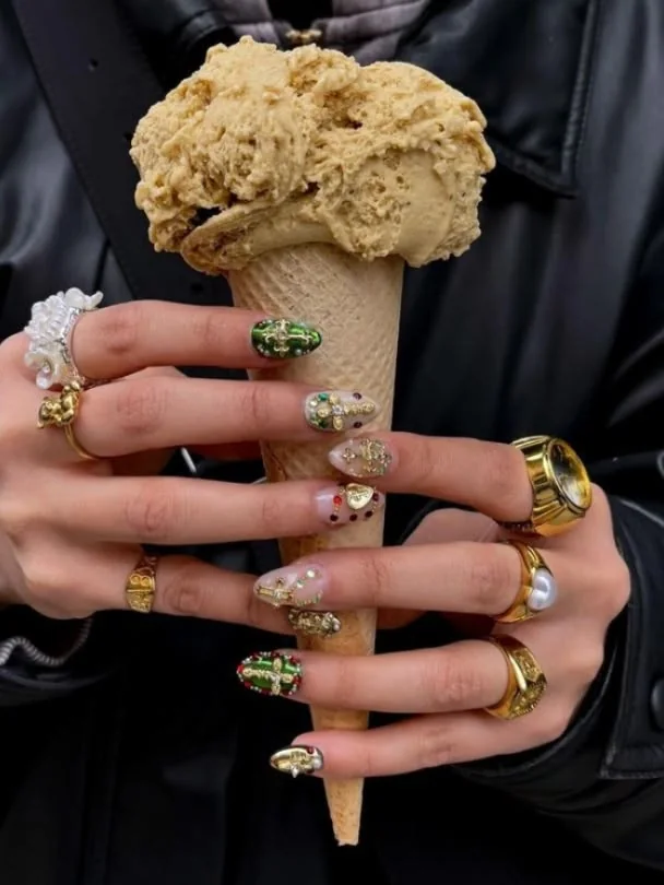 A hand with jewelled and gold-charmed press-on nails holding an ice cream cone