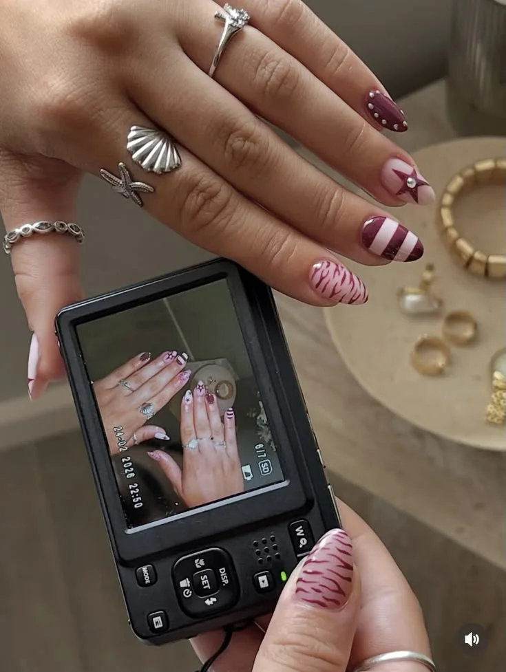 A hand with striped and star nail art holding a digital camera, beside a tray of gold rings