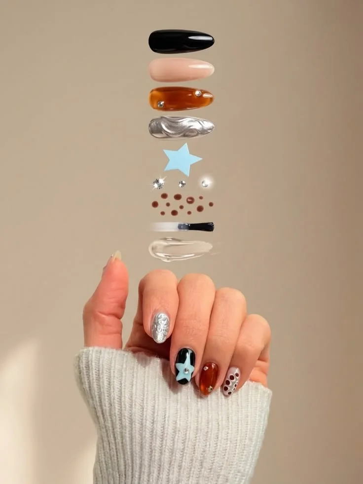 A hand below a vertical stack of separated nail-art components. A black nail, a nude nail, an amber nail, a chrome nail, a blue star, crystals, dots, a brush and clear gel