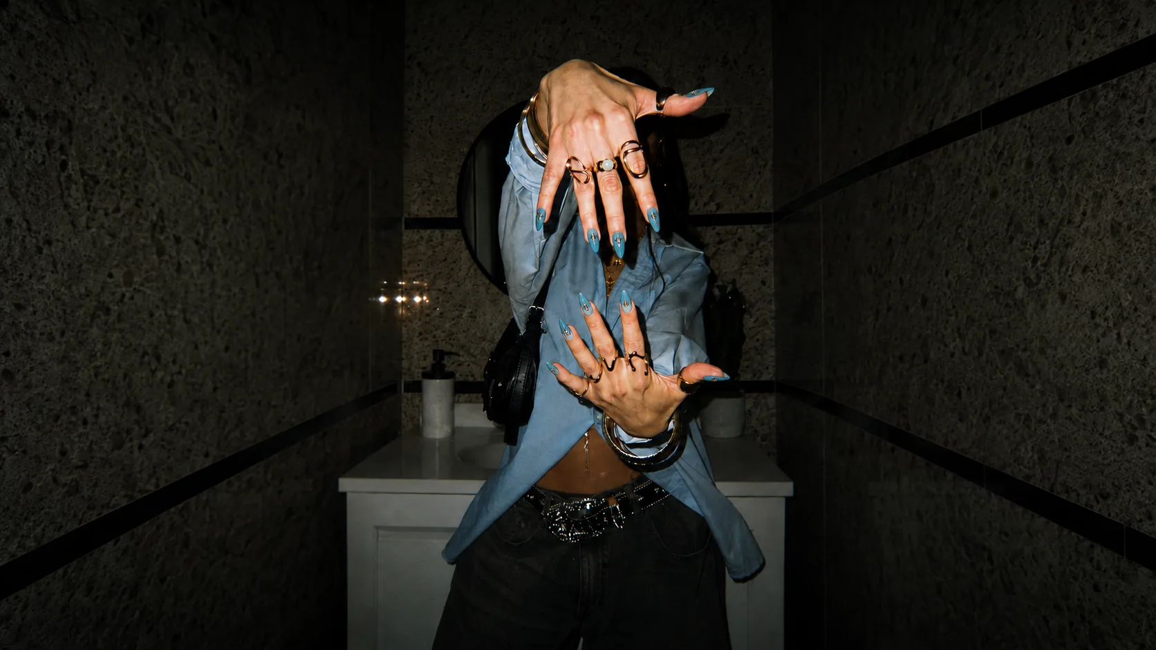 A woman in a blue shirt and stacked gold rings holding both hands to the camera, wearing long ice-blue press-on nails, in a dark stone bathroom