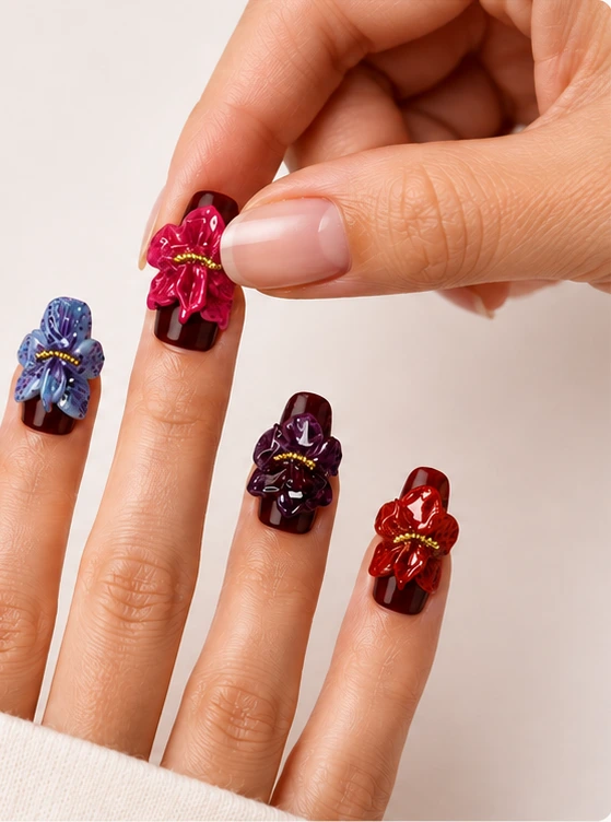 A hand wearing deep burgundy press-on nails with sculpted 3D flowers in blue, purple, magenta and red, while the other hand presses a magenta flower nail into place