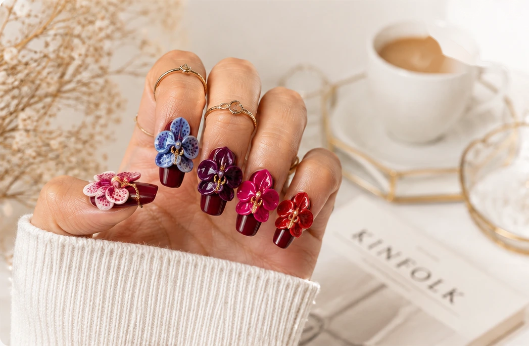 A hand wearing burgundy press-on nails with sculpted orchid flowers and stacked gold rings, resting beside a cup of coffee and a magazine